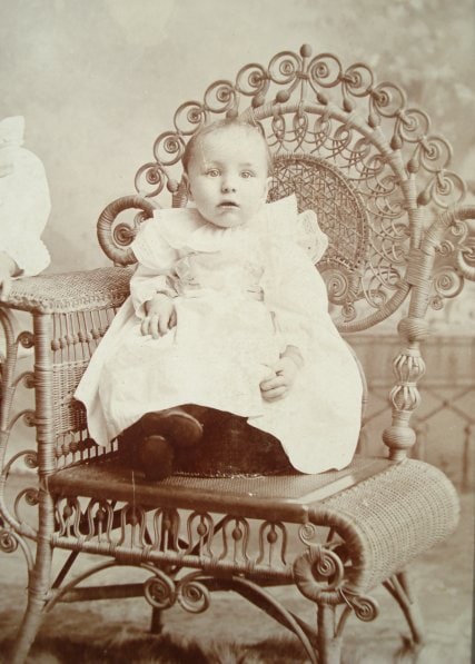 Picture "cabinet card" of a young infant propped on a Victorian wicker photographer's chair, circa 1890s. Part of mother's arm on left side of image. 