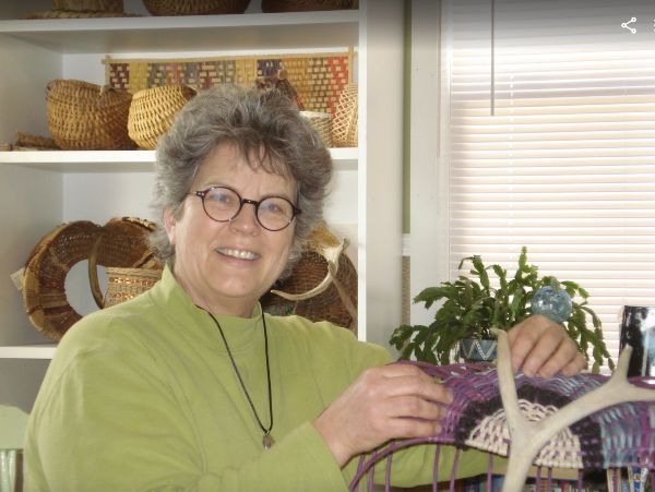 Cathryn Peters, wearing a green shirt, in her studio weaving a large, antler basket in shades of purple. 